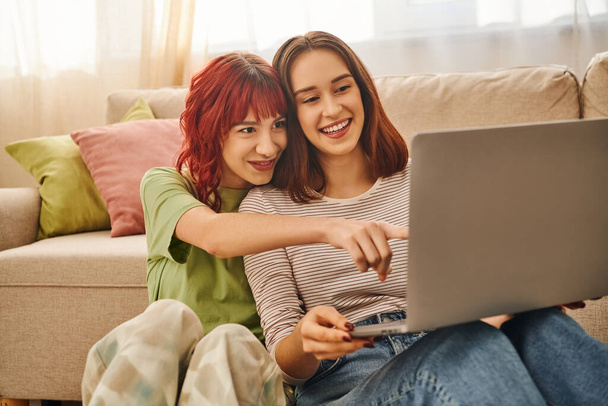 happy moment of cheerful lesbian couple pointing at laptop and watching movie in living room - Foto, afbeelding