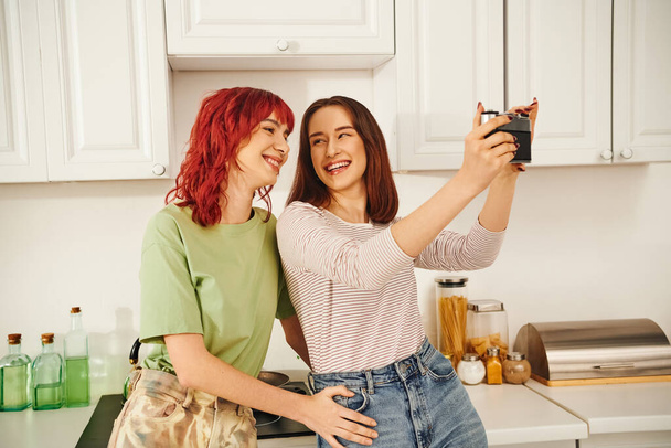 молодая лесбийская пара улыбается и делает селфи на ретро-камеру на кухне, запечатлевая счастливый момент - Фото, изображение