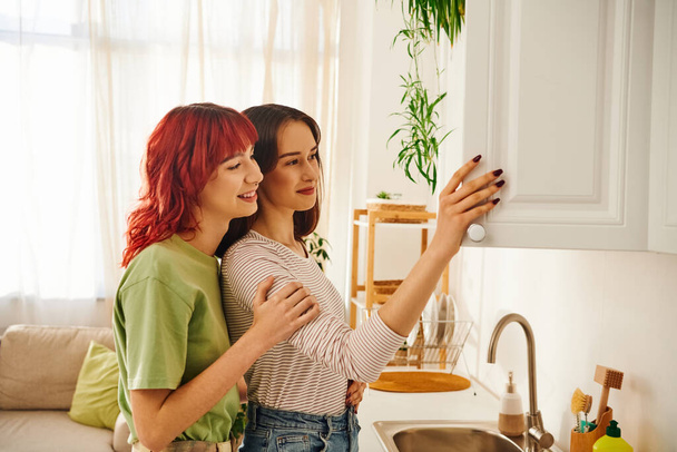 Affectionate young lesbian couple opening white kitchen cabinet and smiling together, domestic life - Photo, Image