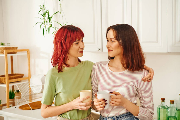 happy lgbt couple sharing a warm beverage while holding cups in their kitchen filled with light - Photo, Image