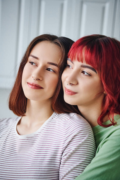 portrait of joyful and pierced lesbian couple looking away and spending time together at home - Photo, Image