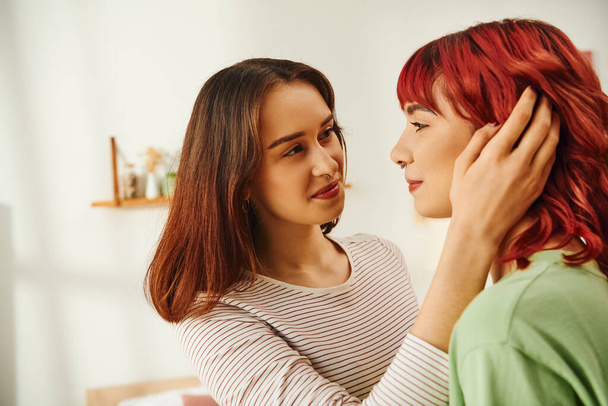 young lesbian woman adjusting red hair of her cheerful girlfriend in modern apartment, bliss - Fotografie, Obrázek