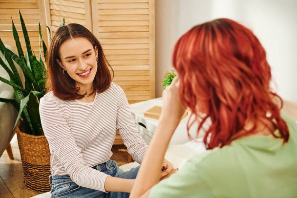 gelukkig jong lesbisch vrouw kijken naar haar vriendin met rood haar en glimlachen in moderne slaapkamer - Foto, afbeelding