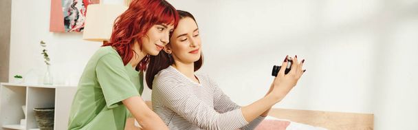 pancarta de sesión de fotos en casa de feliz joven pareja de lesbianas tomando selfie en la cámara retro en el dormitorio - Foto, imagen