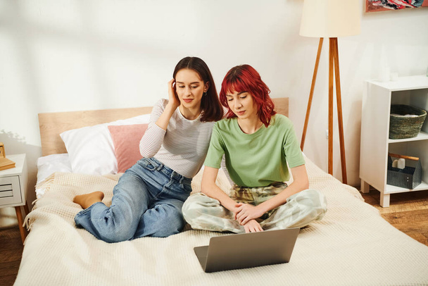 幸せな若いレズビアンカップルでカジュアルな服装で寝室のラップトップでオンラインで映画を見ています - 写真・画像