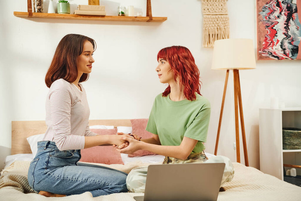 happy young lesbian women holding hands and sitting together on bed next to laptop, lgbt couple - Photo, Image