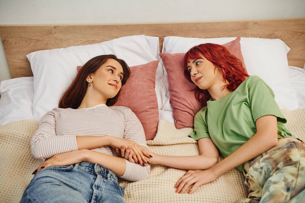 glückliches junges lesbisches Paar, das sich anschaut, während es zusammen auf dem Bett liegt und Händchen hält - Foto, Bild