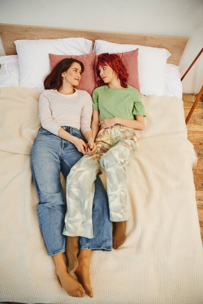 top view of happy lesbian couple holding hands while lying together on bed, looking at each other - Foto, imagen