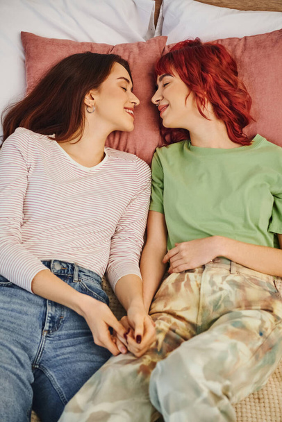 top view of smiling lesbian couple holding hands and lying together on bed, looking at each other - Photo, Image