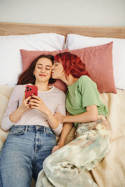 top view of woman with red hair kissing cheek of her girlfriend with smartphone while lying on bed - Photo, Image