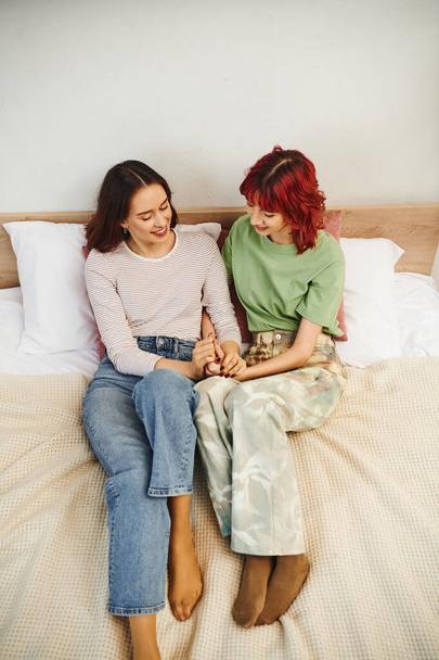 zadowolona młoda lesbijka para w luźnym ubraniu trzymając się za ręce siedząc razem na łóżku - Zdjęcie, obraz