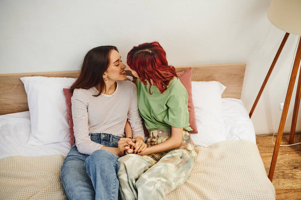 pleased young lesbian couple in casual wear holding hands and going to kiss while sitting on bed - Фото, зображення