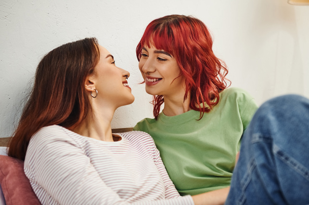 joyful young lesbian couple in casual wear looking at each other while sitting together on bed - Fotó, kép