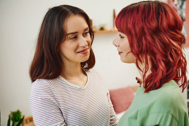 portrait of young and happy lesbian woman looking at her female partner with red hair at home - Фото, изображение