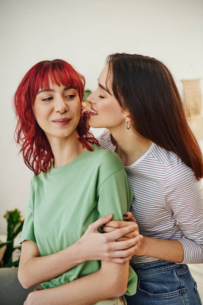 portret młodej i uśmiechniętej lesbijki patrzącej na swoją partnerkę z rudymi włosami w domu - Zdjęcie, obraz