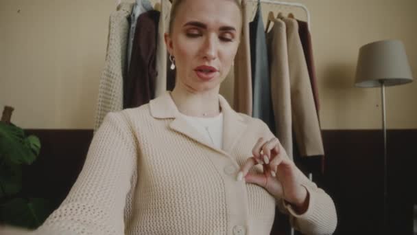若い白人女性ファッションブロガーのハンドヘルドPOVショットは,ミニマリストアパートでソーシャルメディアのビデオを録画しながら,ボタンで加入者にそれを実証するスタイリッシュなニットカーディガンを身に着けています - 映像、動画