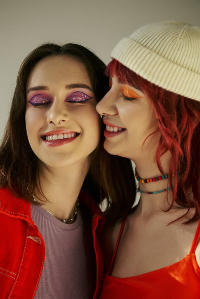 Jeune couple lesbien élégant avec un maquillage audacieux souriant sur fond gris, partageant un moment tendre - Photo, image