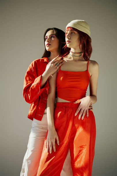 δύο κομψές γυναίκες σε ζωντανή ενδυμασία στέκεται μαζί σε γκρι φόντο, μοντέρνα λεσβιακό ζευγάρι - Φωτογραφία, εικόνα