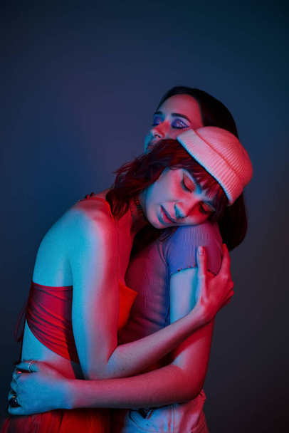 молода лесбійська пара зі сміливим макіяжем і стильним одягом, обіймаючись під фіолетовими вогнями в студії - Фото, зображення