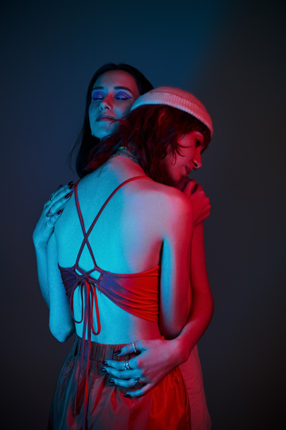 young lgbt couple with bold makeup and stylish attire hugging under purple lights in studio - Photo, Image