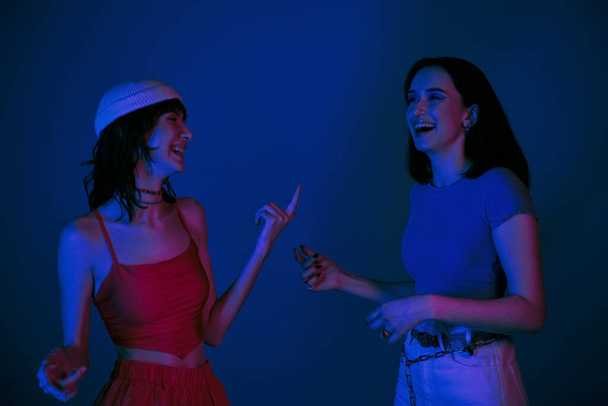 щасливі жінки 20-х років, з сміливим макіяжем і стильним одягом, що сміється і жестикулює під фіолетовими вогнями - Фото, зображення