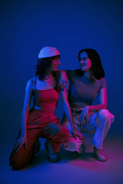 happy girlfriends in 20s posing in stylish attire and looking at each other under purple lights - Photo, Image
