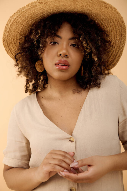 Een mooie jonge Afro-Amerikaanse vrouw met krullend haar met een strohoed en een wit shirt straalt elegantie uit in een studio. - Foto, afbeelding