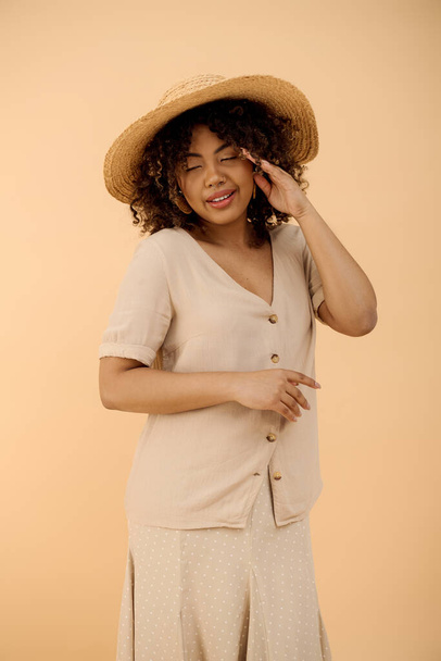 Eine schöne junge Afroamerikanerin mit lockigem Haar, trägt Hut und Kleid und strahlt Eleganz in einem Studio-Ambiente aus. - Foto, Bild