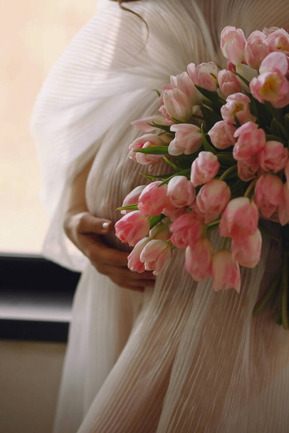Mujer embarazada con un elegante vestido de marfil sosteniendo su vientre con una mano un ramo de tulipanes rosados en la otra mano. Foto de cerca de la barriga embarazada. Concepto de maternidad con estilo. Maternidad feliz - Foto, Imagen