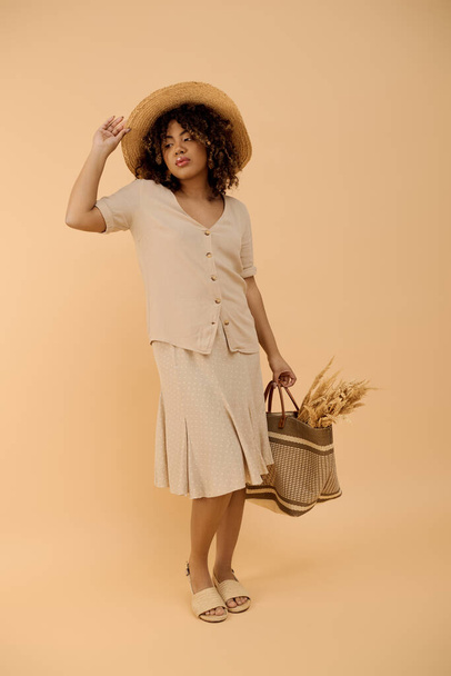 Молодая афроамериканка с вьющимися волосами, в летнем платье и шляпе, элегантно держащая корзину в студии. - Фото, изображение