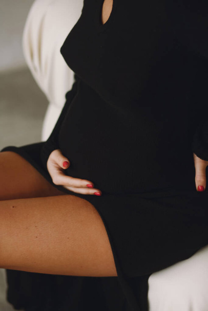 Hamile karnının yakın fotoğrafını çek. Siyah elbiseli hamile bir kadın evdeki sandalyede oturuyor ve hamile karnını tutuyor. Şık annelik kavramı. Metin için alanı kopyala - Fotoğraf, Görsel