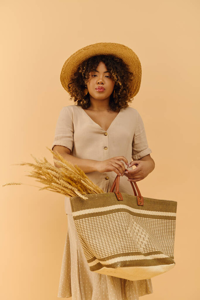 Молода афроамериканка з кучерявим волоссям, одягнена в літню сукню, тримає сумку в солом'яному капелюсі. - Фото, зображення