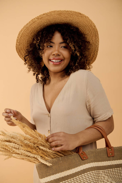 Μια κομψή Αφροαμερικανή γυναίκα με σγουρά μαλλιά, ντυμένη με καλοκαιρινό φόρεμα, κρατώντας μια τσάντα φορώντας ψάθινο καπέλο. - Φωτογραφία, εικόνα
