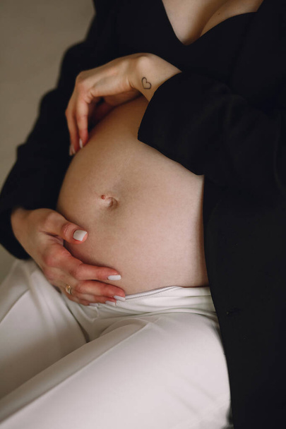 Mujer embarazada con chaqueta negra y pantalones blancos sosteniendo su vientre en el estudio. Concepto de amor. 30 semanas de embarazo. Feliz embarazo. - Foto, Imagen