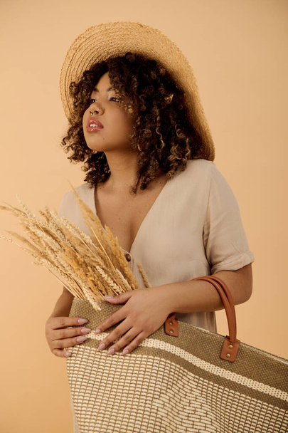 Fiatal afro-amerikai nő göndör hajjal, szalmakalapot visel, táskát tart egy stílusos stúdióban.. - Fotó, kép