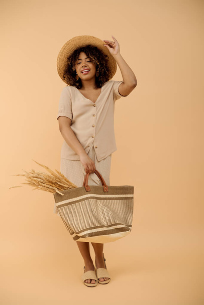Egy gyönyörű, fiatal afro-amerikai nő göndör hajjal, kalapban és ruhában, egy kosarat tart egy stúdióban.. - Fotó, kép