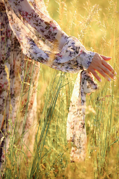 Femme en belle robe de fleur touchant et agitant l'herbe haute tout en marchant dans la prairie. Toucher à la main l'herbe sauvage sur le terrain. Photo en gros plan - Photo, image