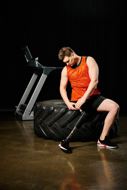 Ένας άνδρας με ένα προσθετικό πόδι κάθεται πάνω από ένα μαύρο λάστιχο σε ένα γυμναστήριο, επιδεικνύοντας δύναμη και αποφασιστικότητα στη ρουτίνα προπόνηση του. - Φωτογραφία, εικόνα