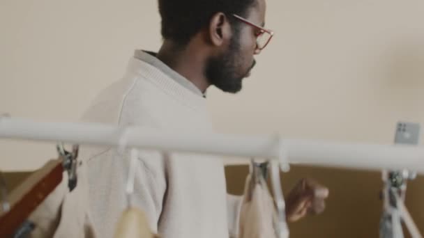Slowmo selbstbewusster junger männlicher schwarzer Modeblogger und Stylist demonstriert trendige Artikel auf Kleiderbügeln, während er Lehrvideos für seinen Kanal auf einem Stativ filmt - Filmmaterial, Video