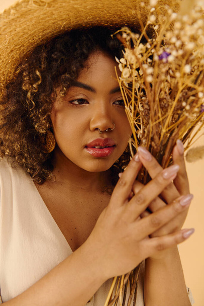 Kaunis nuori afroamerikkalainen nainen olkihattu päässä pitäen joukko elinvoimainen kukkia kihara tukka studiossa. - Valokuva, kuva