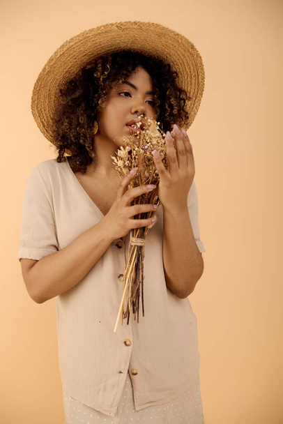 ストロー帽子をかぶったカーリーヘアの若いアフリカ系アメリカ人女性,スタジオの設定で活気に満ちた花束を保持. - 写真・画像