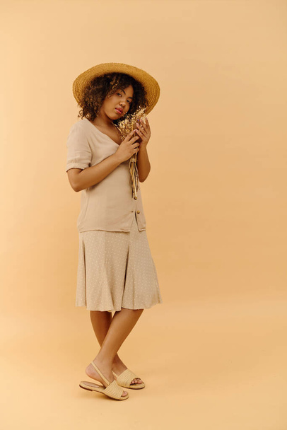 Una bella giovane donna afroamericana con i capelli ricci tiene un fiore mentre indossa un cappello alla moda in un ambiente da studio. - Foto, immagini