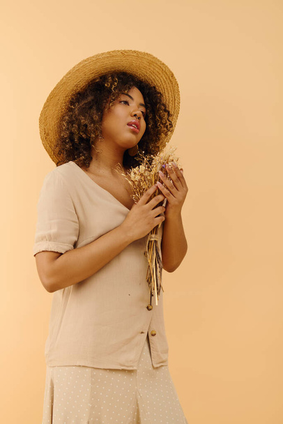 Uma bela jovem afro-americana com cabelo encaracolado usando um chapéu de palha, segurando flores secas em uma pose pacífica. - Foto, Imagem