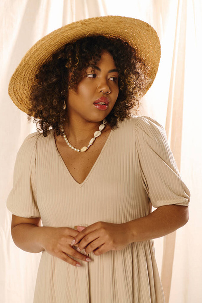 Eine schöne junge Afroamerikanerin mit lockigem Haar posiert mit Strohhut und Sommerkleid in einem Studio-Setting. - Foto, Bild
