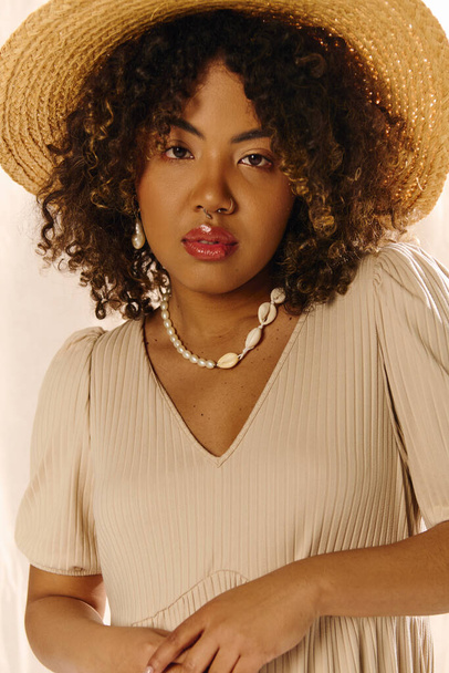 Μια όμορφη νεαρή Αφροαμερικανή γυναίκα με σγουρά μαλλιά είναι κομψά ντυμένη με καλοκαιρινό φόρεμα και ψάθινο καπέλο. - Φωτογραφία, εικόνα