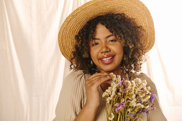 Eine junge Afroamerikanerin mit lockigem Haar, trägt einen Strohhut und hält in einem Atelier einen lebendigen Blumenstrauß in der Hand. - Foto, Bild