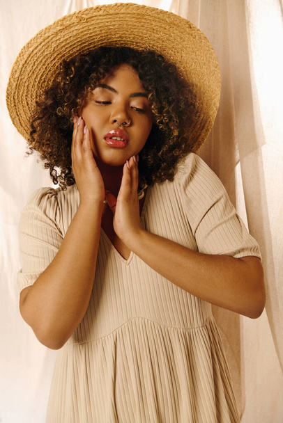 Una hermosa joven afroamericana con el pelo rizado usando un sombrero de paja, sosteniendo sus manos a la cara en una pose serena. - Foto, Imagen