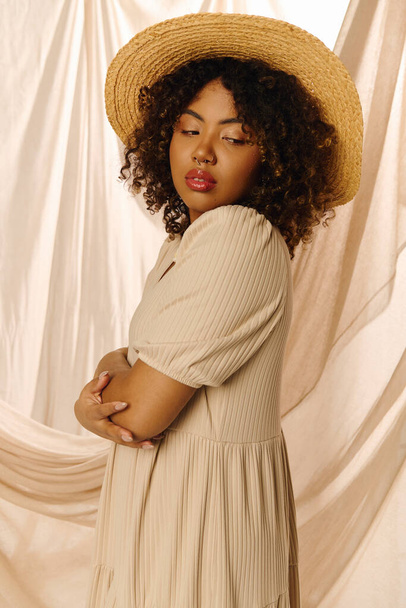 Eine schöne junge Afroamerikanerin mit lockigem Haar posiert im Sommerkleid und Strohhut in einem Studio-Setting. - Foto, Bild