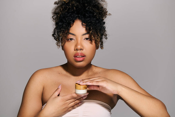 Egy fiatal afro-amerikai nő, göndör hajjal, törölközőbe csomagolva, egy üveg tejszínt tart egy stúdióban.. - Fotó, kép