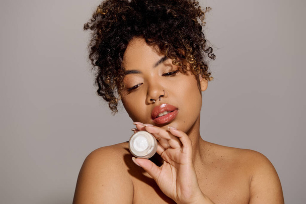 Joven mujer afroamericana con el pelo rizado sosteniendo tarro de crema en su cara para la rutina de cuidado de la piel en un entorno de estudio. - Foto, imagen